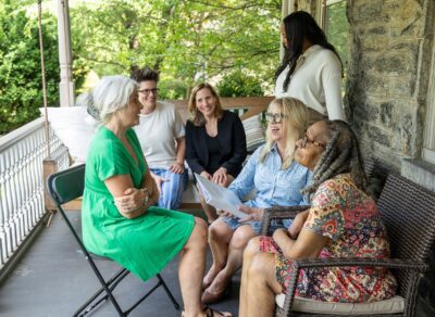 A group of people sitting on a porch talking and laughing, focused on an older adult.